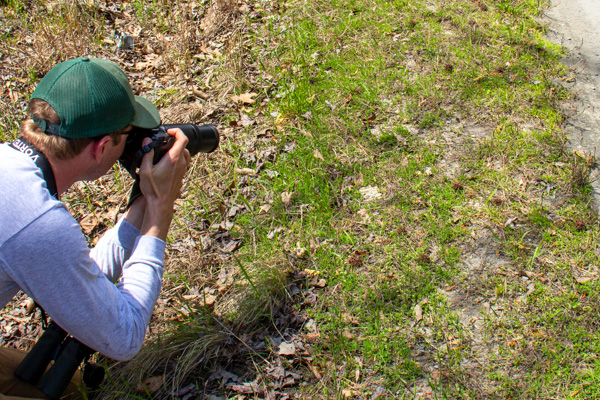 Ảnh chụp người đội mũ xanh đang dùng máy ảnh chụp cảnh ven đường có cỏ mọc thưa thớt.